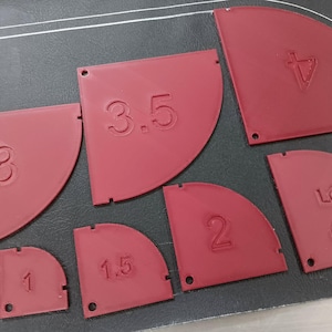 May include: A collection of red, semi-circular quilting templates with numbers from 1/2 to 4. Each template has a small hole and is arranged on a dark surface. The numbers are embossed on the templates.