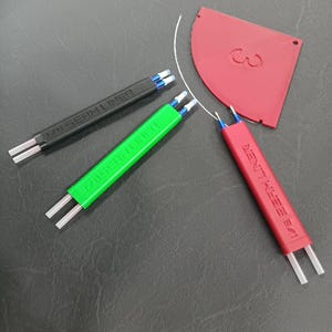 May include: An overhead shot shows three seam liners in black, green, and red, along with a red plastic tool. The seam liners have the text "1/4 SEAM LINER" printed on them. The red tool has the number "3" cut out of it.