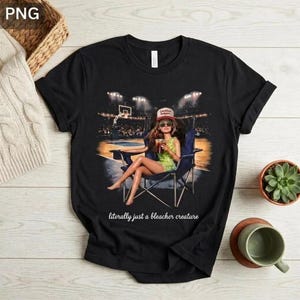 May include: Black t-shirt featuring a graphic of a person in a folding chair at a basketball game, holding a drink. The person is wearing a hat and sunglasses. Text at the bottom reads "literally just a bleacher creature."