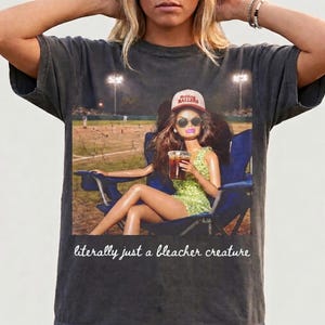 Peut inclure: T-shirt gris foncé avec un imprimé d'une poupée dans un stade de baseball, portant des lunettes de soleil et un chapeau avec l'inscription "Holding Ballers". La poupée tient une boisson et est assise dans une chaise bleue. Le texte "literally just a bleacher creature" est en dessous.