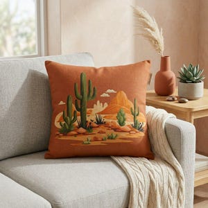May include: An orange decorative pillow with a desert landscape design. The pillow features green cacti, a tan mountain, and a blue sky. The pillow is square and rests on a gray sofa.