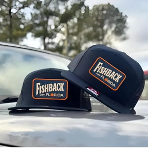 May include: Two navy blue trucker hats with a rectangular patch featuring the text "FISHBACK FOR FLORIDA" in white and orange. The hats have a mesh back and a flat brim.