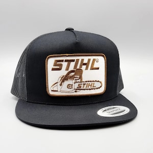 May include: Black trucker hat with a mesh back and a flat brim. The front features a rectangular patch with the brand name "STIHL" and a chainsaw graphic in brown and white. The hat has a snapback closure.