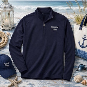 May include: A navy blue long-sleeve quarter-zip pullover featuring the text Coastal Vibe embroidered in white on the left chest. The garment has a stand-up collar, a silver-toned zipper pull, and raglan sleeves, providing a clean and simple look.