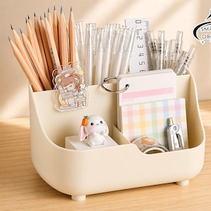 Op de afbeelding: Een crèmekleurige bureau-organizer gevuld met potloden, pennen, notitieblokken en briefpapier. Inclusief een klein konijnenbeeldje en een doorzichtige sticker. De organizer heeft meerdere compartimenten.