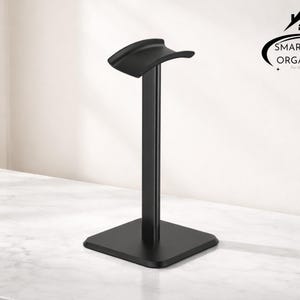 May include: A black headphone stand with a curved top and a square base. The stand is made of metal and is designed to hold headphones. The Smartspace Organizer logo is in the upper right corner.