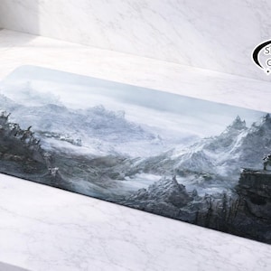 May include: Rectangular desk mat depicting a mountainous landscape in shades of grey and white, with a figure on a cliff. The mat has a logo in the top right corner. The design evokes a sense of adventure.