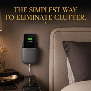 Op de afbeelding: Een donkergrijze wandhouder voor telefoons met een zwarte smartphone met een groen batterijpictogram en de tekst 100% CHARGED. Boven de houder staat de tekst THE SIMPLEST WAY TO ELIMINATE CLUTTER in gouden letters op een donkere muur.