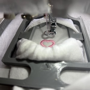 May include: Close-up of an embroidery machine in operation, stitching a pink heart design onto white fabric. The machine's needle is poised above the fabric, with a grey frame holding the material in place. The focus is on the detailed stitching process.