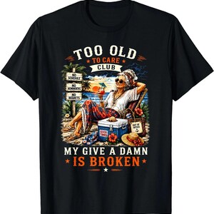 May include: A black t-shirt featuring a graphic of a person relaxing in a beach chair with a drink. Text reads: TOO OLD TO CARE CLUB, NO SCHEDULE, NO REMINDERS, NO REGRETS, GOOD TIMES, OUT OF OFFICE, MY GIVE A DAMN IS BROKEN. The scene includes a cooler and palm trees.