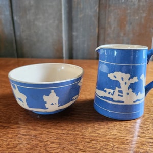 May include: A blue and white ceramic sugar bowl and creamer set with a white silhouette design. The design features a horse and rider, a tree, and a fence.