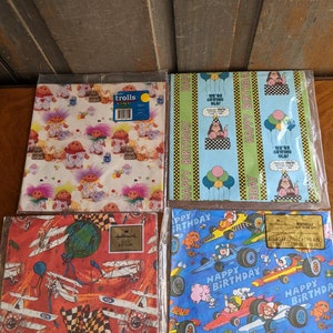 Pu&ograve; includere: Quattro rotoli di carta da regalo con diversi design. Il primo rotolo presenta un motivo colorato di troll. Il secondo rotolo ha uno sfondo blu con un motivo ripetitivo di palloncini e il testo "Buon compleanno". Il terzo rotolo ha uno sfondo rosso con un motivo di aerei e il testo "Buon compleanno". Il quarto rotolo ha uno sfondo blu con un motivo di auto da corsa e il testo "Buon compleanno".