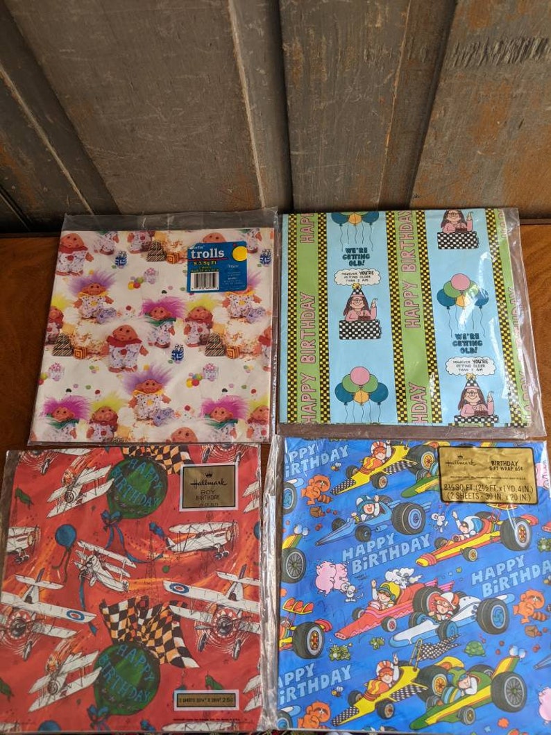 Pu&ograve; includere: Quattro rotoli di carta da regalo con diversi design. Il primo rotolo presenta un motivo colorato di troll. Il secondo rotolo ha uno sfondo blu con un motivo ripetitivo di palloncini e il testo "Buon compleanno". Il terzo rotolo ha uno sfondo rosso con un motivo di aerei e il testo "Buon compleanno". Il quarto rotolo ha uno sfondo blu con un motivo di auto da corsa e il testo "Buon compleanno".