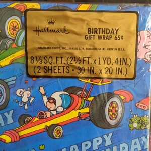 Pu&ograve; includere: Una carta da regalo di compleanno blu e gialla con un'auto da corsa dei cartoni animati con un pilota che indossa un casco e occhiali protettivi. La carta include anche un topo, rane e il testo "Buon compleanno".