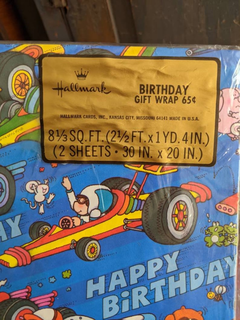 Pu&ograve; includere: Una carta da regalo di compleanno blu e gialla con un'auto da corsa dei cartoni animati con un pilota che indossa un casco e occhiali protettivi. La carta include anche un topo, rane e il testo "Buon compleanno".