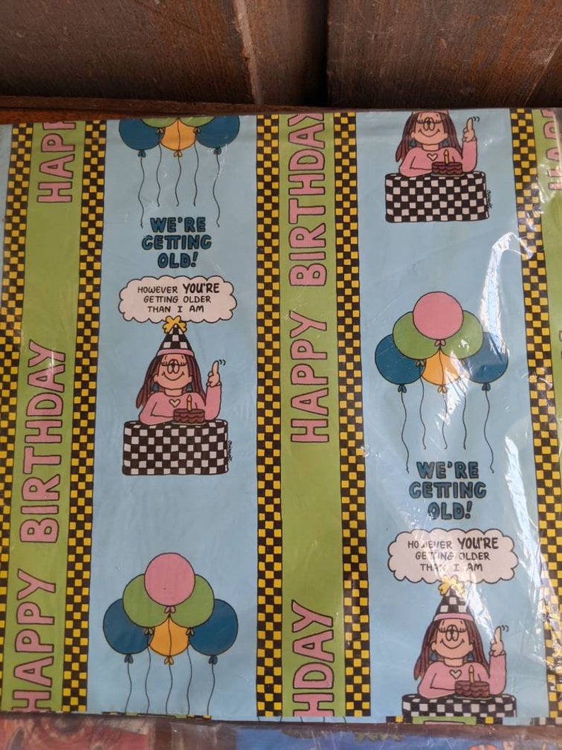 Pu&ograve; includere: Carta da regalo blu e verde con un motivo a scacchi e strisce gialle. La carta presenta un personaggio dei cartoni animati con un vestito rosa e un cappello di compleanno. Il testo sulla carta recita "We're getting old! However you're getting older than I am."