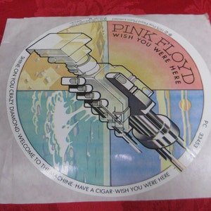 May include: A round sticker with a pink, blue, and yellow background. The sticker features a robotic arm reaching out from the center. The text "Pink Floyd Wish You Were Here" is printed in black on the pink background. The text "Shine On You Crazy Diamond Welcome To The Machine Have A Cigar Wish You Were Here" is printed in black around the edge of the sticker.