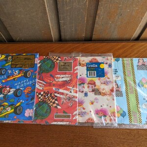 Pu&ograve; includere: Quattro diversi design di carta da regalo con una variet&agrave; di temi. Il primo design &egrave; blu con auto da corsa e le parole "Buon compleanno". Il secondo design &egrave; rosso e bianco con aerei e le parole "Buon compleanno". Il terzo design &egrave; rosa e bianco con troll e le parole "Trolls". Il quarto design &egrave; blu e verde con palloncini e le parole "Buon compleanno".
