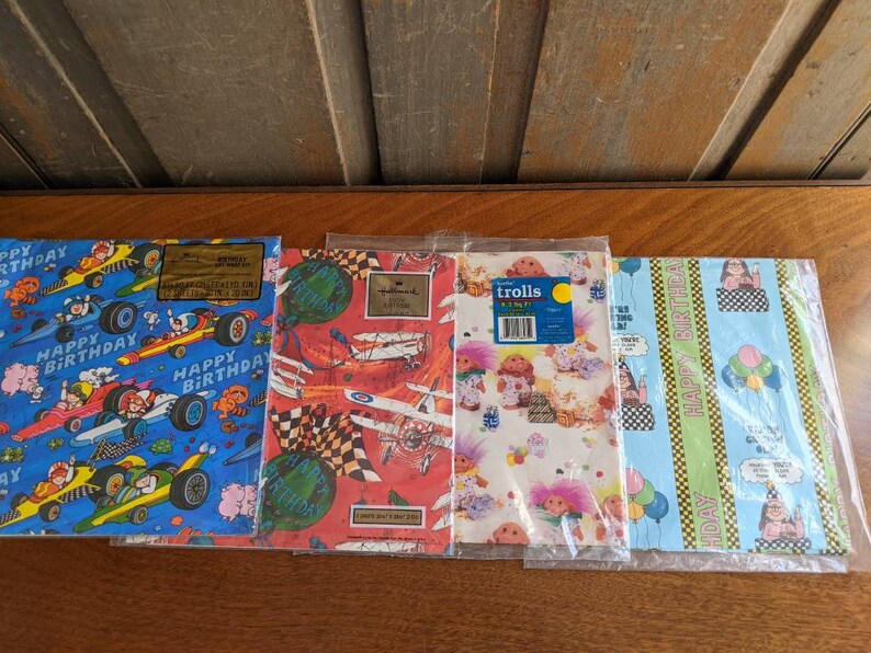 Pu&ograve; includere: Quattro diversi design di carta da regalo con una variet&agrave; di temi. Il primo design &egrave; blu con auto da corsa e le parole "Buon compleanno". Il secondo design &egrave; rosso e bianco con aerei e le parole "Buon compleanno". Il terzo design &egrave; rosa e bianco con troll e le parole "Trolls". Il quarto design &egrave; blu e verde con palloncini e le parole "Buon compleanno".