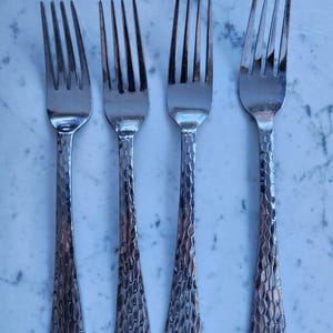 May include: Four stainless steel forks with textured handles. The tines are straight and evenly spaced. The handles have a hammered design, adding a rustic touch to the silverware. The forks are arranged in a row.