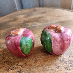 May include: Two ceramic apple-shaped salt and pepper shakers. The shakers are primarily red with green leaf accents and brown stems. The shakers are sitting on a wooden surface.