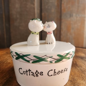 May include: A white ceramic cottage cheese container with a lid featuring a green and black design. Two kissing cat figurines with pink and green accents sit atop the lid. The words "Cottage Cheese" are written in black script on the container.