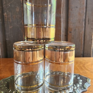 May include: Set of three clear glass tumblers with gold trim. The tumblers have a gold band around the top and a gold band around the bottom. The gold band in the middle is a pattern of vertical lines.