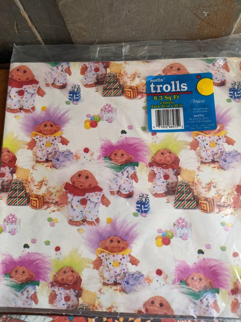 Pu&ograve; includere: Carta da regalo con un motivo ripetuto di troll colorati con cappelli festivi e coriandoli. La carta &egrave; bianca con uno sfondo blu chiaro. Il testo sull'etichetta dice "norfin trolls 8.3 Sq Ft 2 sheets Each 20 in x 30 in Artfaire This giftwrap is recyclable norfin Total E.P.S. 1.8KG &copy; 1992 Troll Company 71922 66575 2"
