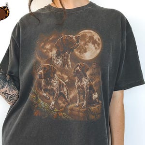 Può includere: T-shirt grigio scuro con una grafica di tre Pointer tedeschi sotto la luna piena. I cani sono marroni e bianchi, con uno sfondo di scena di caccia. La maglietta ha un aspetto vintage e usurato.