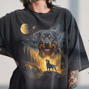 Könnte beinhalten: Schwarzes T-Shirt mit einem Rottweiler-Hundedesign. Die Grafik zeigt ein großes Hundeporträt, einen kleineren Hund auf einem Felsen, einen Mond und eine Waldszene. Das Shirt besteht aus einem weichen, dunklen Stoff.