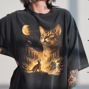 May include: Dark gray t-shirt featuring a detailed graphic of a cat looking up at the moon, with a smaller cat howling in a forest scene. The artwork is rendered in warm tones of brown, gold, and orange.