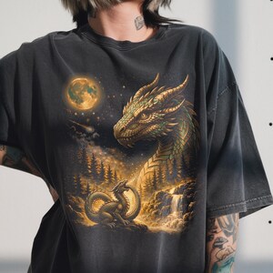 May include: Dark grey t-shirt with a fantasy dragon illustration. The design shows two dragons, a full moon, and a waterfall in a forest setting. The artwork uses gold, green, and brown tones. The t-shirt is a relaxed fit.