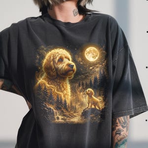 Puede incluir: Camiseta gris oscuro con una ilustración de un perro golden doodle. El diseño incluye una luna llena, árboles y una cascada. La obra de arte tiene una calidad onírica y etérea, con acentos brillantes.