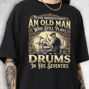 May include: Black t-shirt with a graphic that reads "Never Underestimate An Old Man Who Still Plays Drums In His Seventies." The graphic features a drummer playing a drum set in a vintage style.