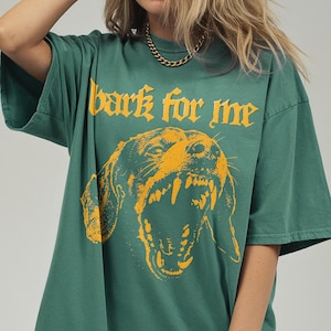 May include: Green t-shirt with the text "bark for me" above a yellow illustration of a snarling dog. The shirt has short sleeves and a relaxed fit. The graphic design is bold and eye-catching.