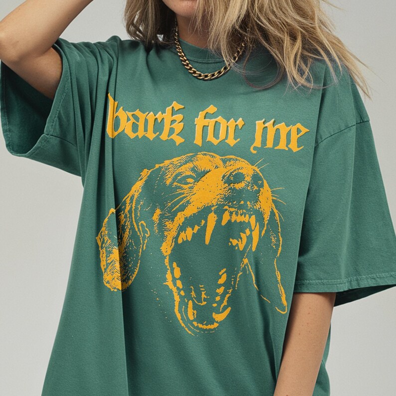 May include: Green t-shirt with the text "bark for me" above a yellow illustration of a snarling dog. The shirt has short sleeves and a relaxed fit. The graphic design is bold and eye-catching.