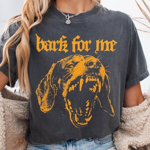 May include: Dark gray t-shirt with the text "bark for me" in orange gothic lettering. Below the text is an orange illustration of a snarling dog's face, showcasing its teeth. The shirt has a vintage, worn look.