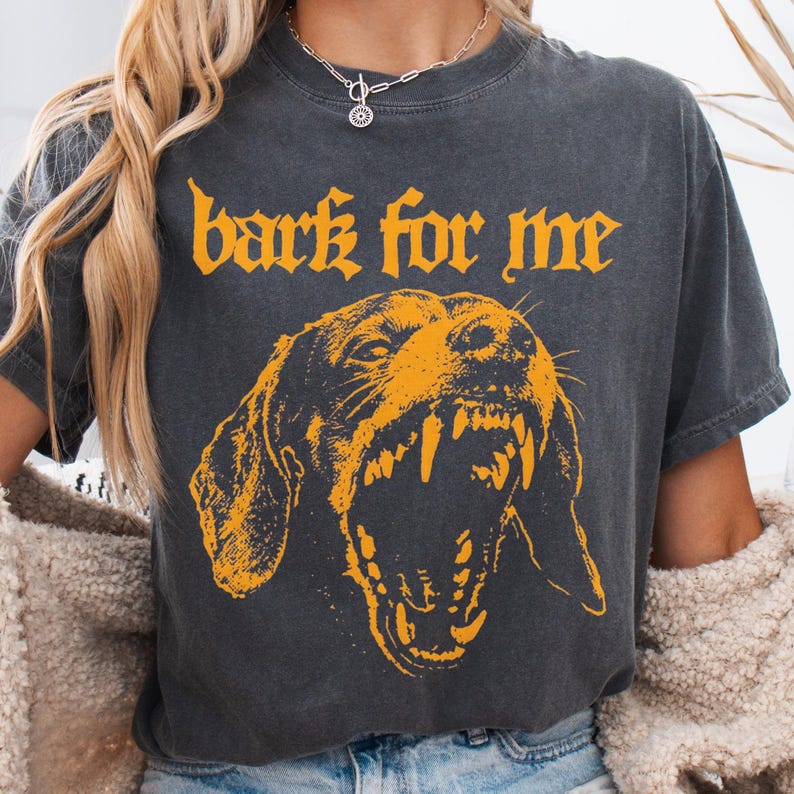 May include: Dark gray t-shirt with the text "bark for me" in orange gothic lettering. Below the text is an orange illustration of a snarling dog's face, showcasing its teeth. The shirt has a vintage, worn look.