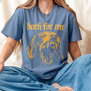 May include: A blue t-shirt with the text "bark for me" in yellow gothic lettering above a yellow illustration of a snarling dog. The shirt has short sleeves and a crew neck.