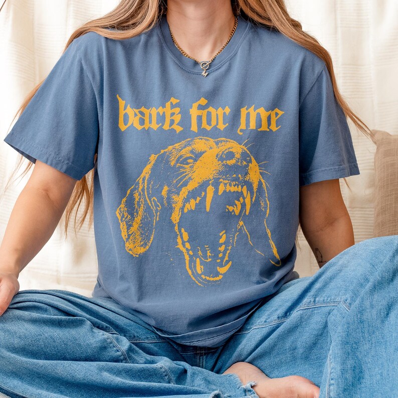 May include: A blue t-shirt with the text "bark for me" in yellow gothic lettering above a yellow illustration of a snarling dog. The shirt has short sleeves and a crew neck.