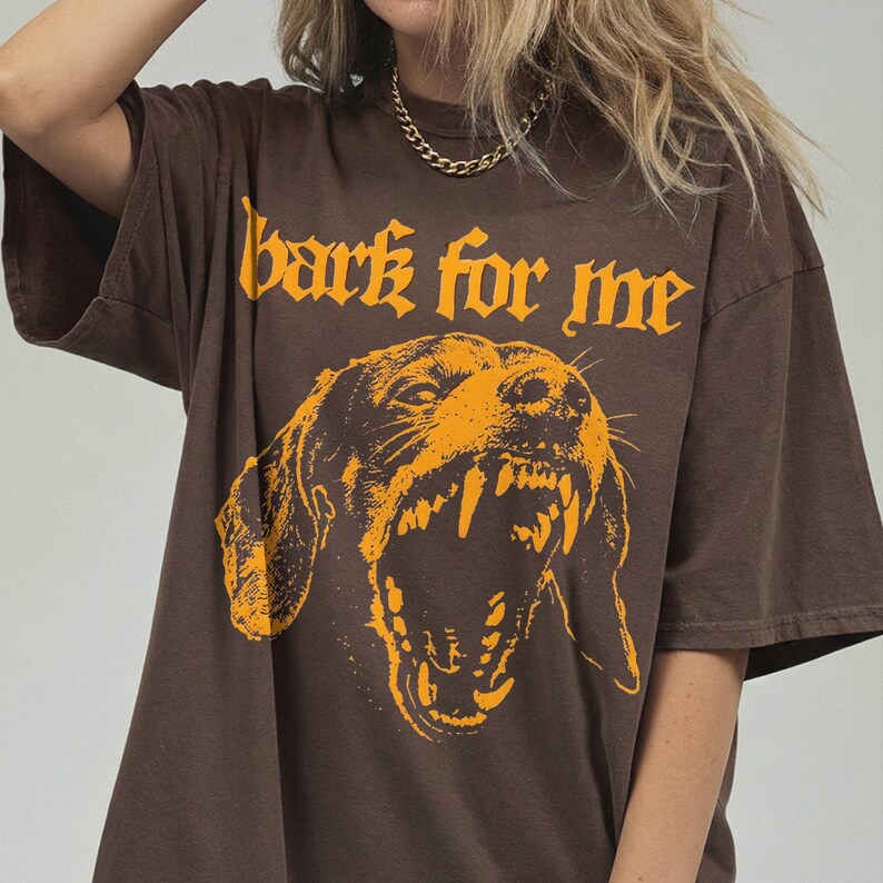May include: Brown t-shirt with the text "bark for me" in orange gothic lettering above an orange illustration of a snarling dog. The shirt has short sleeves and a relaxed fit.