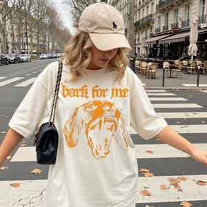May include: Cream-colored t-shirt with the text "bark for me" in orange gothic lettering and a matching orange illustration of a dog's face. The person is wearing a beige baseball cap with a black logo.