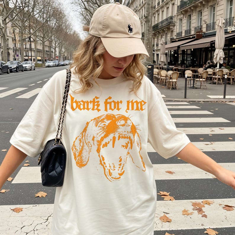May include: Cream-colored t-shirt with the text "bark for me" in orange gothic lettering and a matching orange illustration of a dog's face. The person is wearing a beige baseball cap with a black logo.