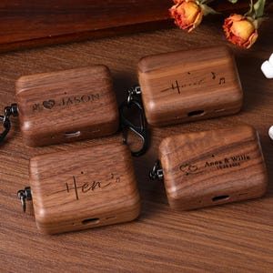 May include: Four personalized wooden AirPod cases with black carabiner clips. Each case is engraved with a name or names, and some include dates and symbols. Two white AirPod earbuds are also visible.
