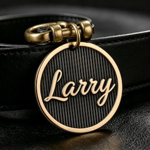 May include: A circular black and gold metal tag featuring the name Larry in elegant gold script lettering. The tag has a vertically striped black background and is attached to a black leather strap by a gold-toned metal ring and hardware.