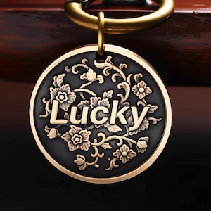 May include: A round, brass-colored pet tag with the word "Lucky" in the center, surrounded by floral and vine designs. The tag has a dark background and a gold-colored border, with a matching ring for attachment.