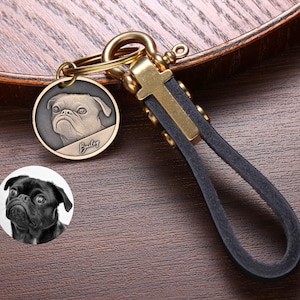 May include: A brass keyring with a circular pendant showing a pug's portrait and the name "Bailey." The keyring has a black leather strap and a gold-coloured clasp. The background is a wooden surface.