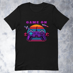 Puede incluir: Camiseta negra con un diseño retro de videojuego. El gráfico presenta un mando de juego morado, una puesta de sol y las palabras "GAME ON" en morado. El diseño incluye acentos azules, naranjas y rosas.