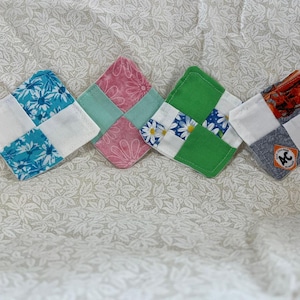 May include: Four handmade square coasters with a patchwork design. Each coaster features a different color scheme: blue floral, pink floral, green with white and blue flowers, and a gray and orange design with an "AC" logo.