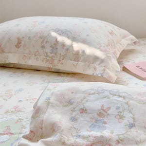 Pode incluir: Conjunto de almofada e lençol creme com estampa floral e de coelho. O design apresenta flores rosa, azuis e verdes e coelhos em um padrão caprichoso. A roupa de cama é feita de um tecido macio e leve.